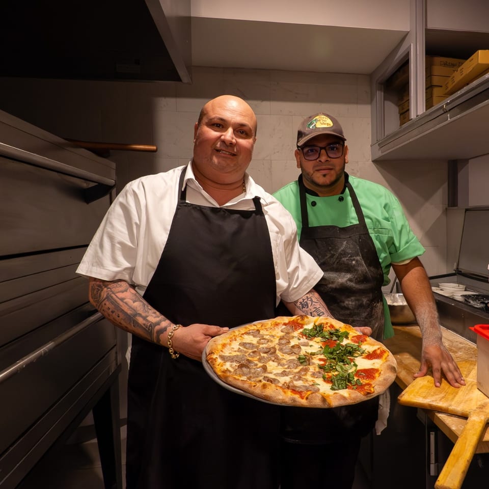 Two Baci Trattoria & Pizza chefs in aprons holding a half-sausage, half-basil pizza in a kitchen.
