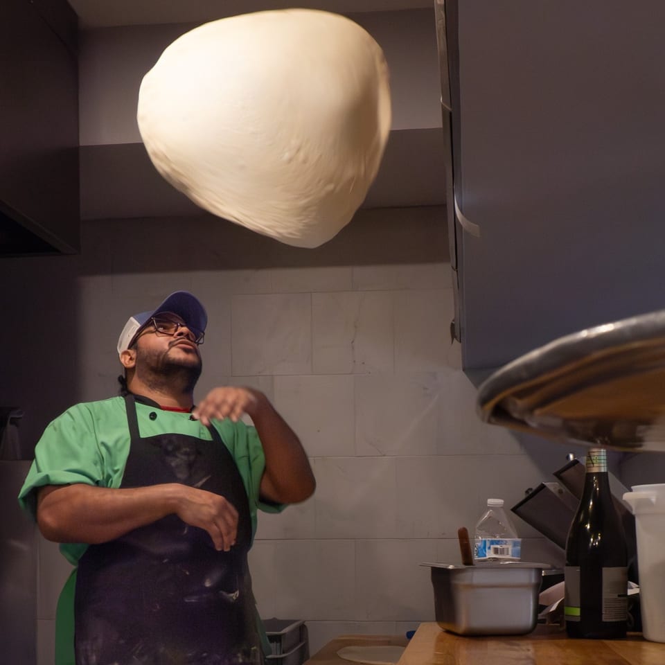 A pizza chef in a green shirt and black apron tossing pizza dough high in the air.