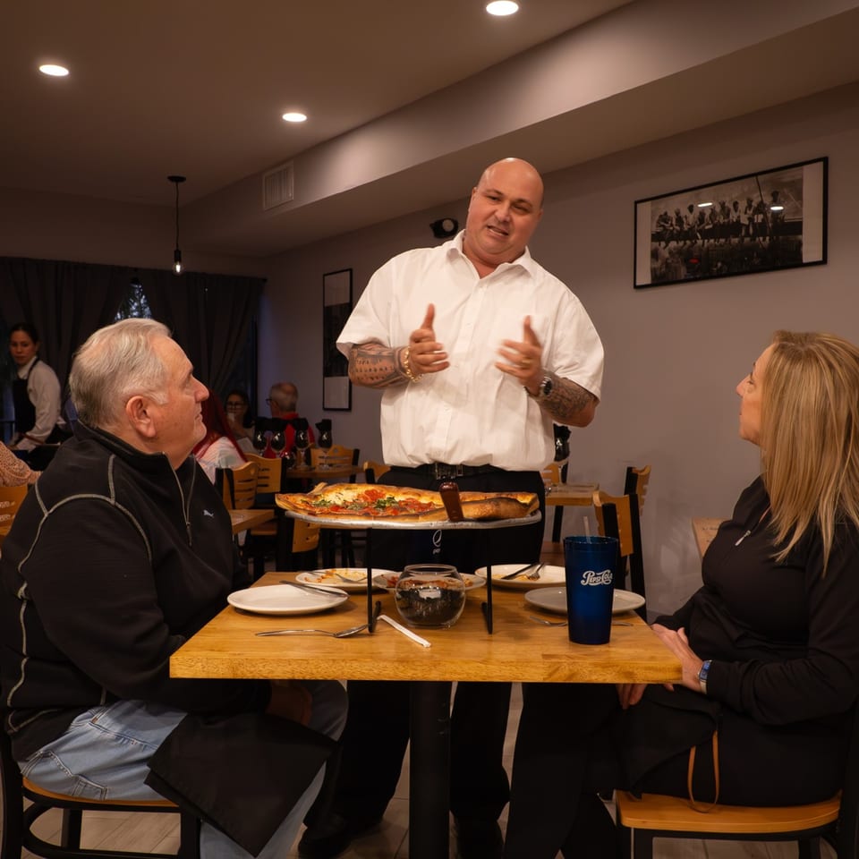 A server at Baci Trattoria & Pizza presents a large pizza to a seated couple.